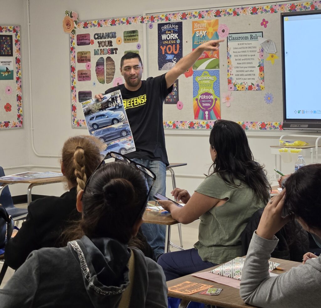 A VC Defensa volunteer in a Defensa shirt presenting to a classroom, pointing at a screen while people at desks follow along with printed handouts.