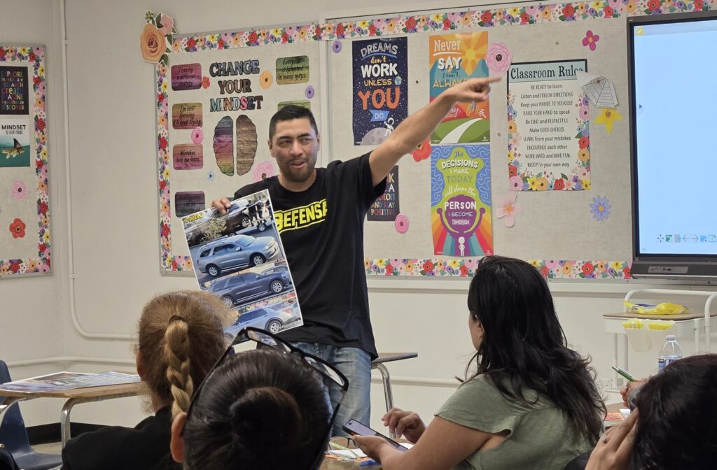 A VC Defensa volunteer in a Defensa shirt presenting to a classroom, pointing at a screen while people at desks follow along with printed handouts.