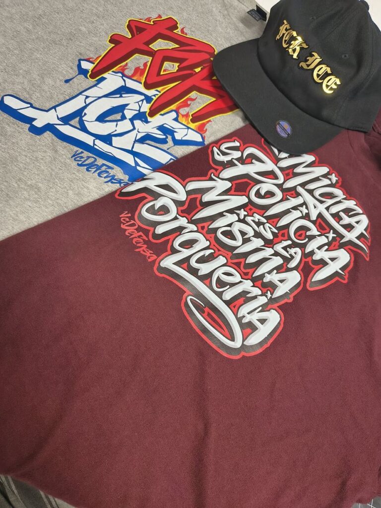 A gray T-shirt with a red, blue and yellow graffiti design, a maroon sweatshirt with white and red graffiti lettering in Spanish, and a black cap with gold lettering, laid flat.